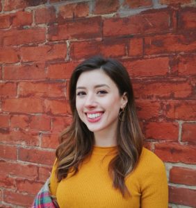 Alexandra Caprara stands in front of a brick wall wearing a yellow shirt and smiling at the camera.