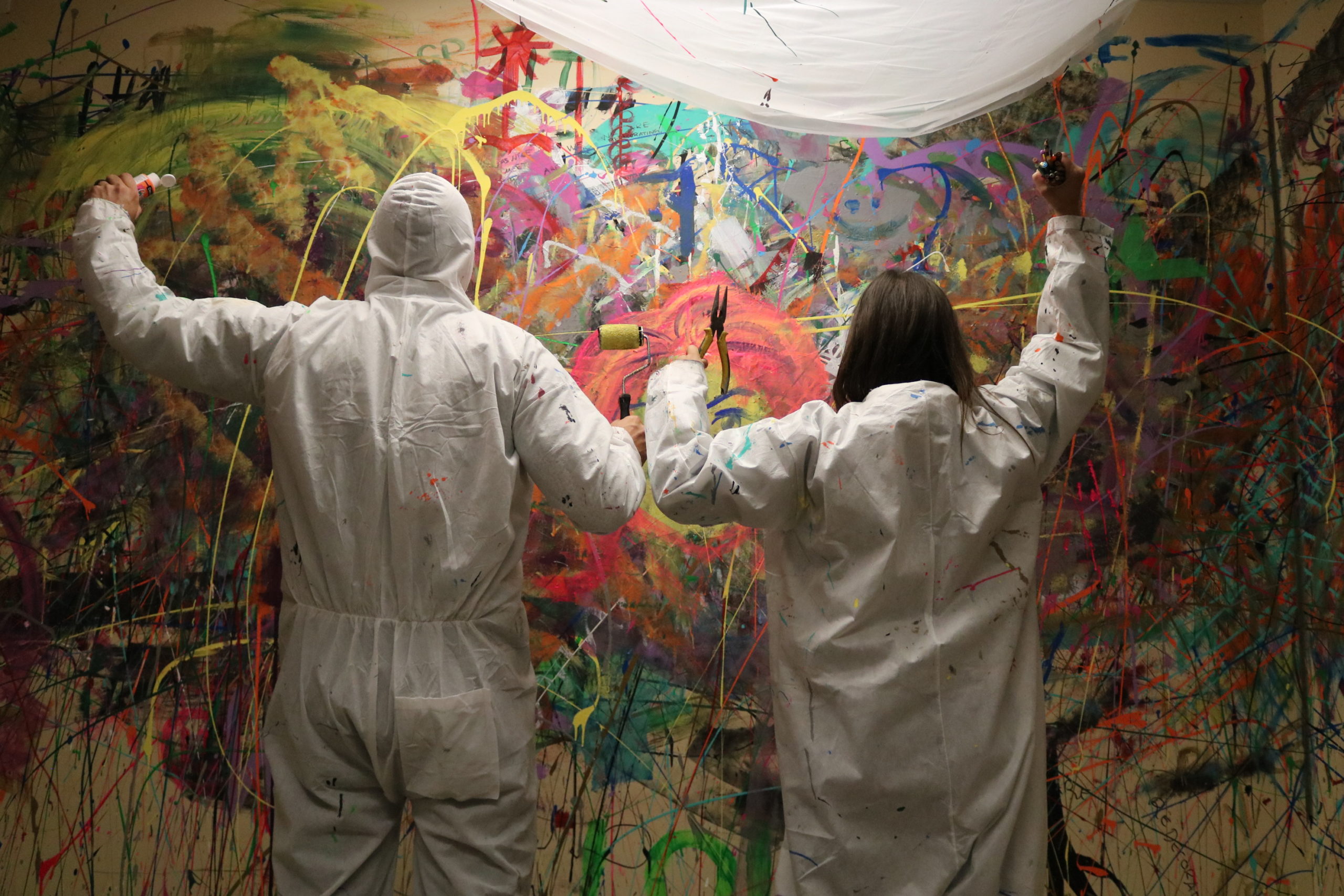 Two people in white painters overalls stand in front of a paint splattered wall.