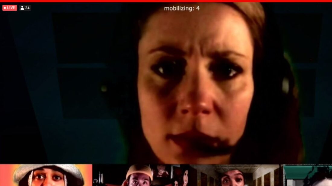 Screen shot from performance of Orestes. A woman wearing a gaming headset with attached microphone looks at the screen in a video chat window. The top portion of four other people in the video chat are visible across the bottom of the image.