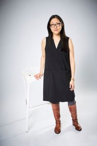 Taien Ng-Chan stands next to a white stool in a black dress, brown boots and glasses.