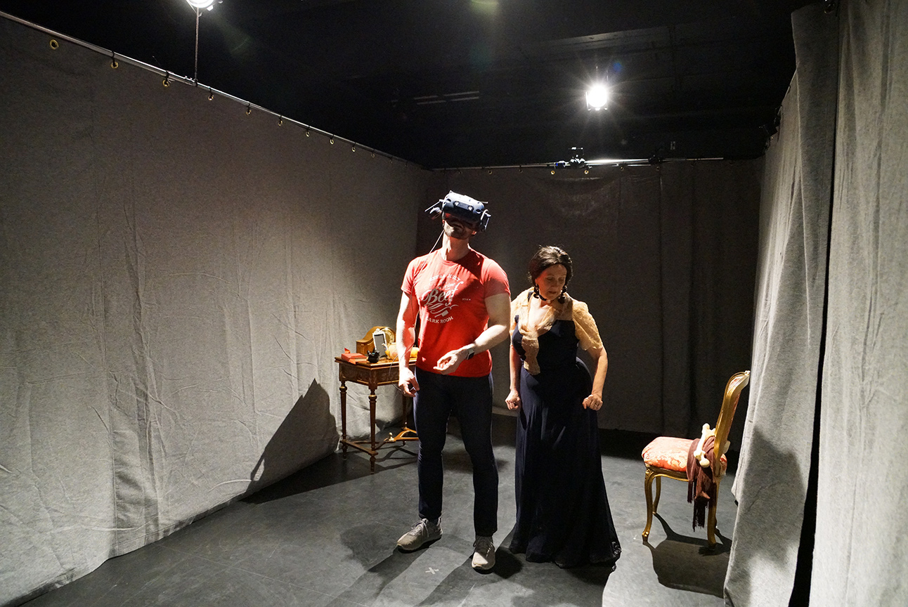 A performer in costume stands behind the audience member wearing a VR headset during a performance of Bury the Wren.