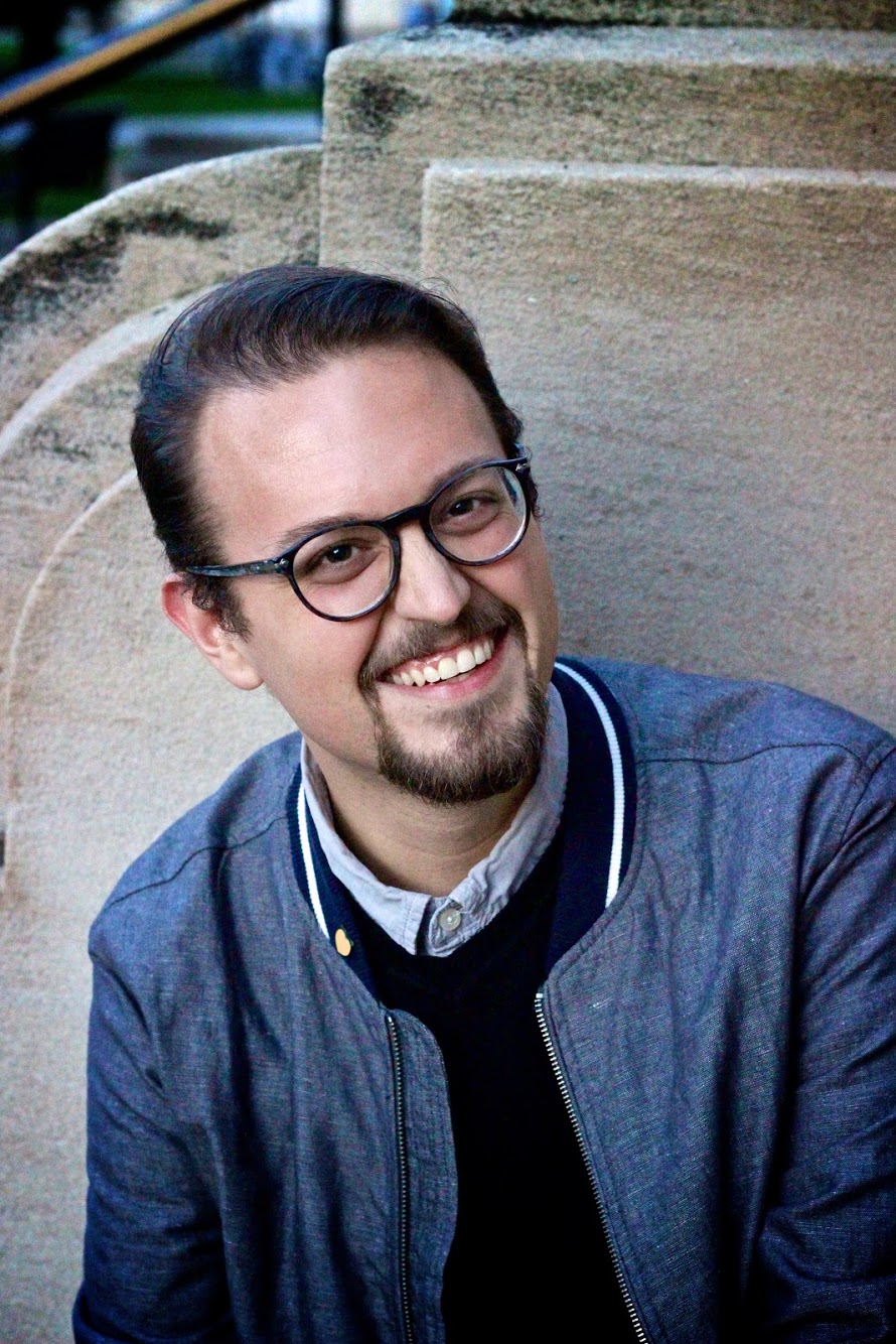 Zachary Scalzo looks at the camera and smiles, wearing a blue jacket and glasses.