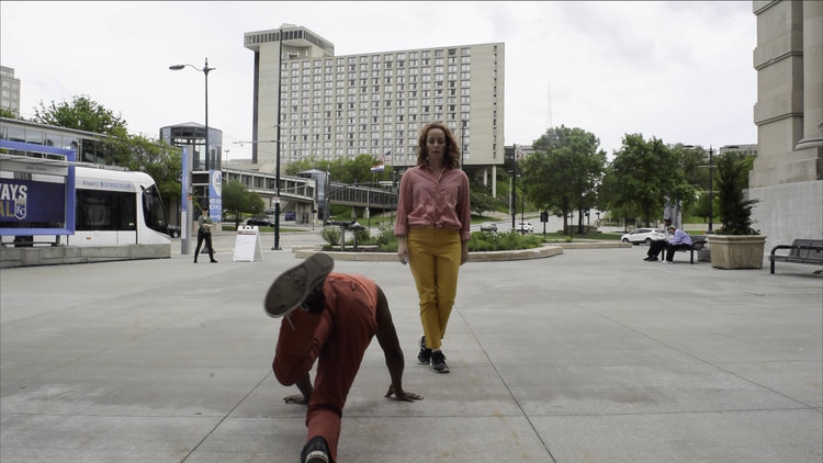 Dancers in colourful street clothes perform on a public sidewalk near a street car stop in Kansas City.